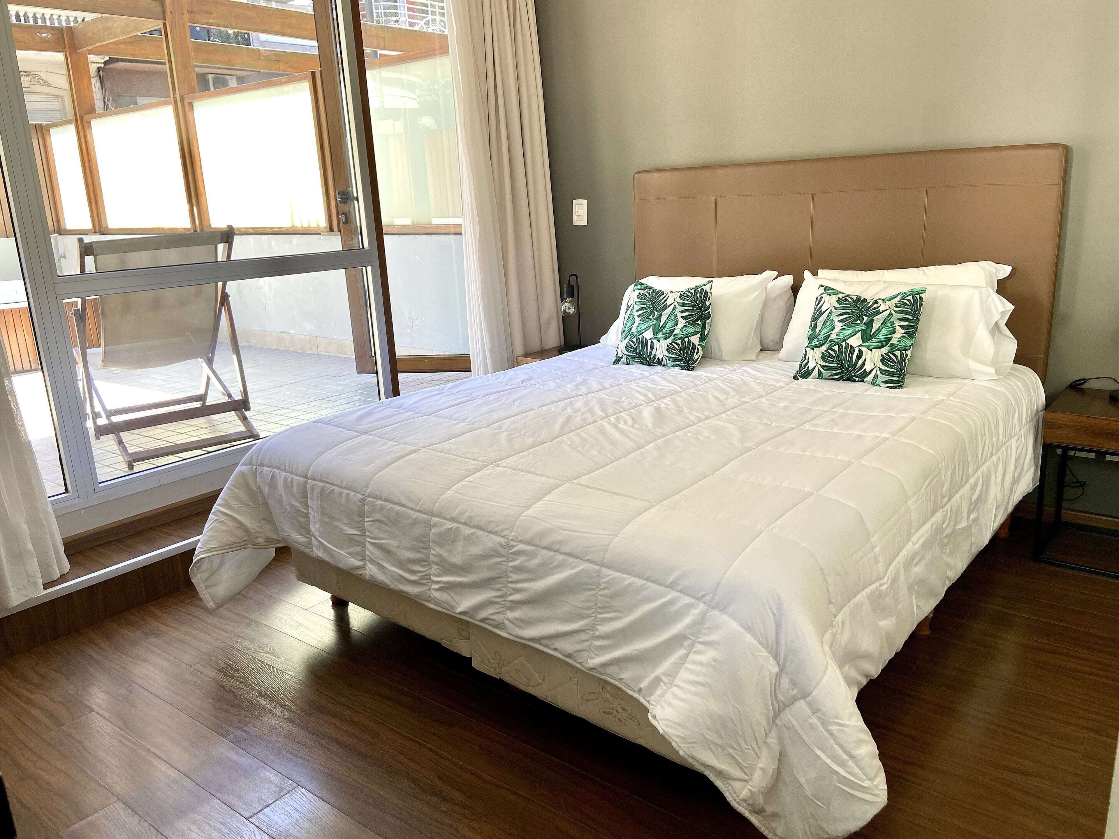A bedroom with a large bed, wooden flooring, a window with curtains, and a view of the outside.