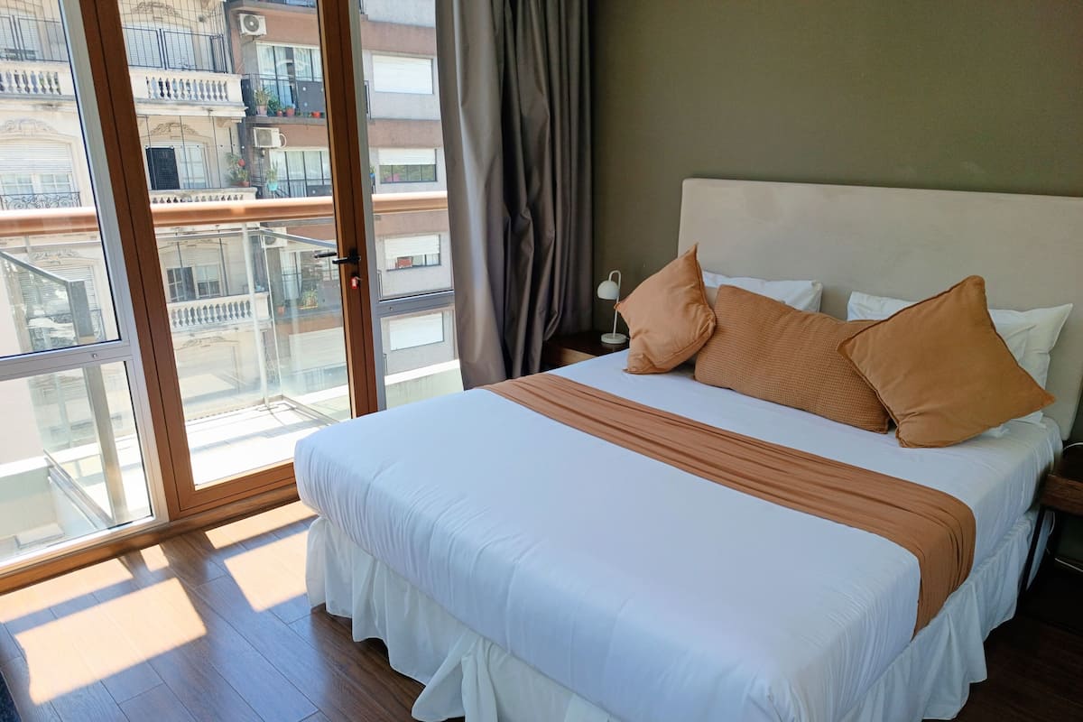 Hotelkamer met een groot bed, nachtkastje, lamp en stadszicht door een glazen deur.