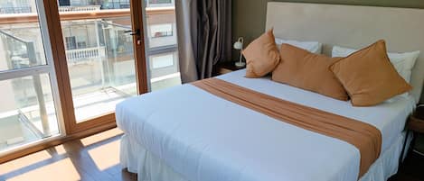 A hotel room with a large bed, a nightstand, a lamp, and a view of the city through a glass door.