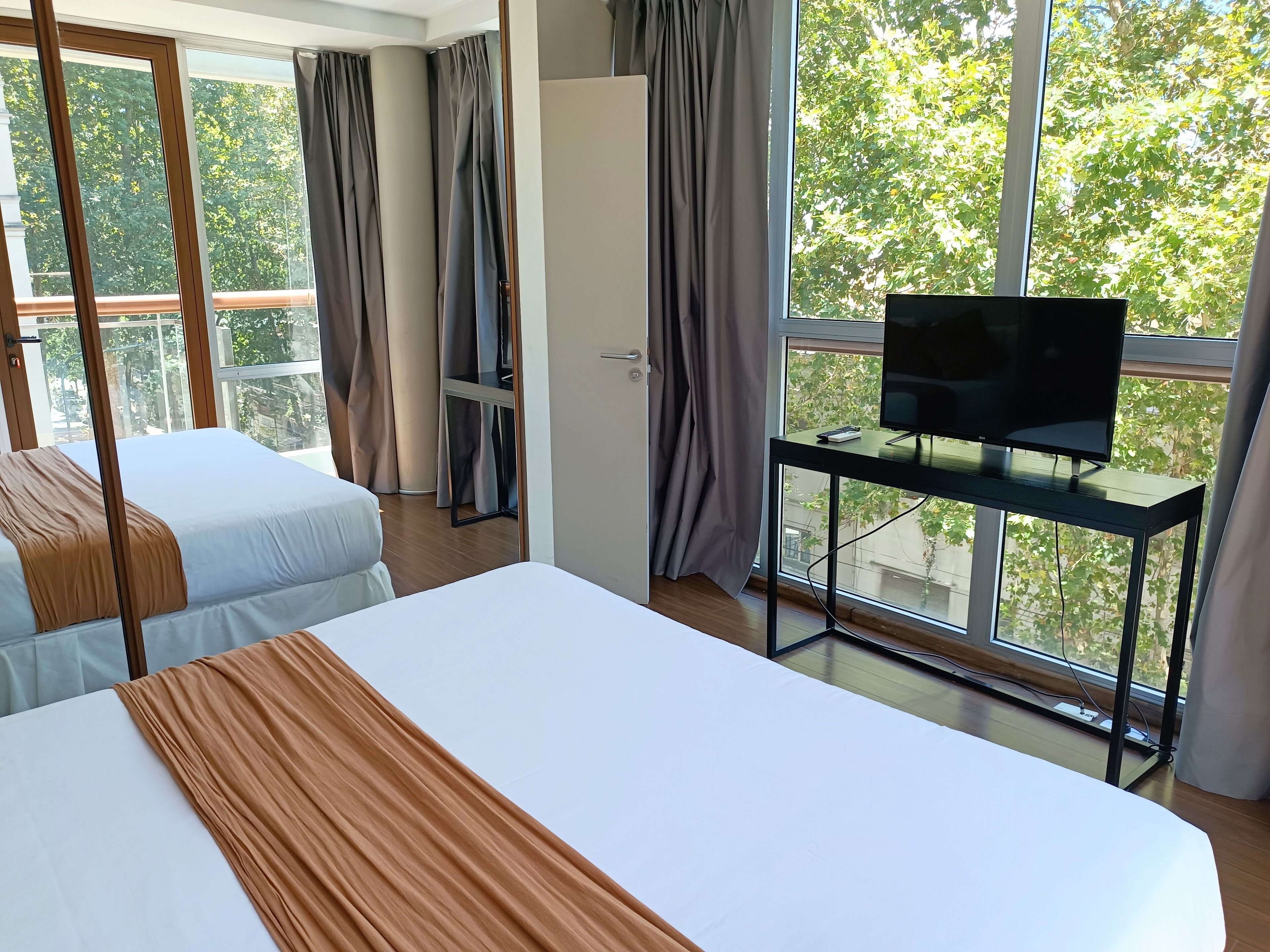 Une chambre d’hôtel avec deux lits, un téléviseur posé sur un meuble et de grandes fenêtres avec des rideaux.