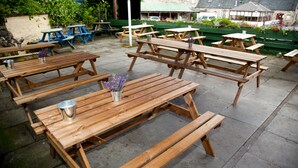 An outdoor patio area with multiple wooden picnic tables and benches, arranged on a paved surface. - Richmond House Hotel (Fort Augustus)