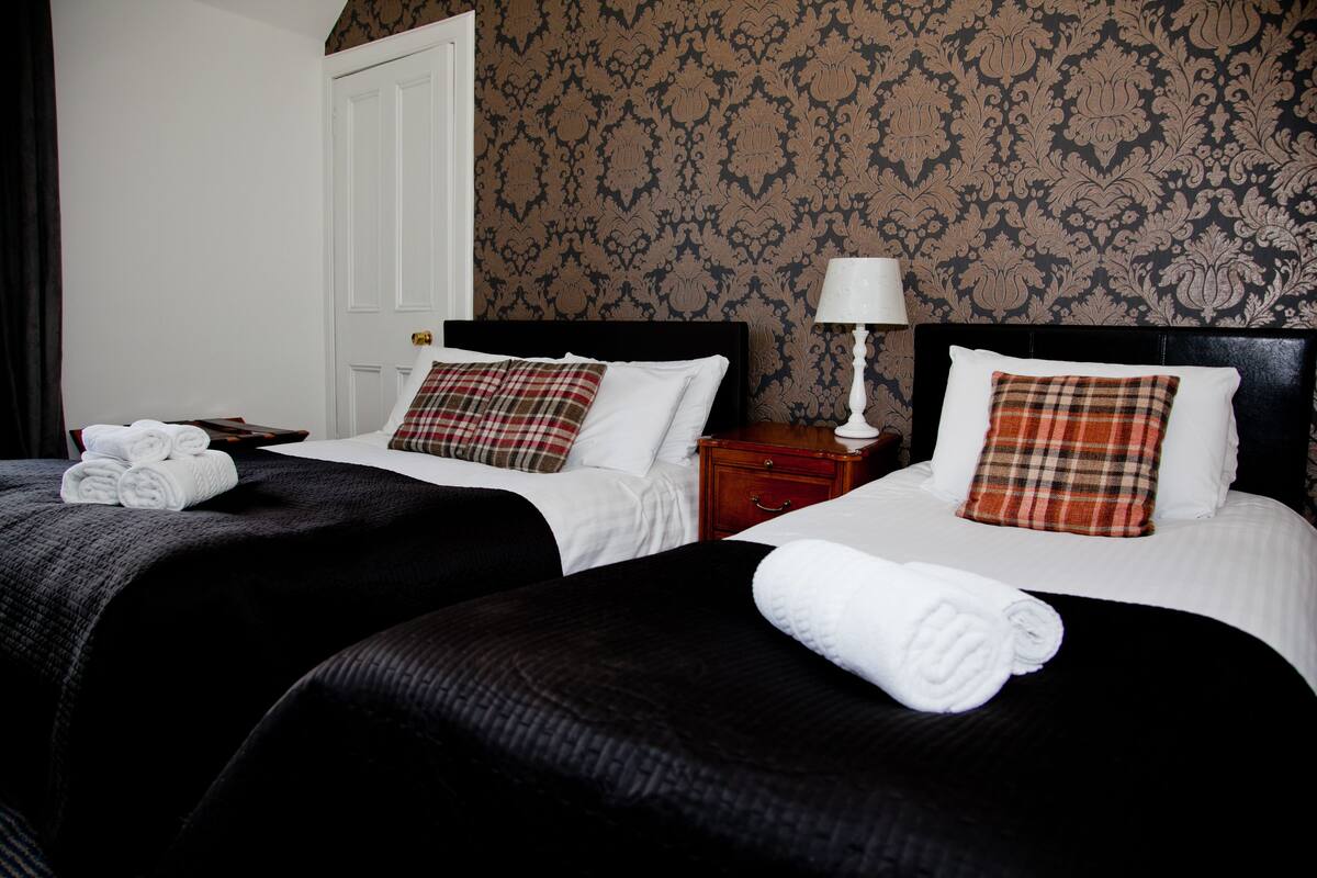 A hotel room with two beds, each with a black headboard, white bedding, and plaid pillows. There is a wooden nightstand with a lamp between the beds, and a door to the left.