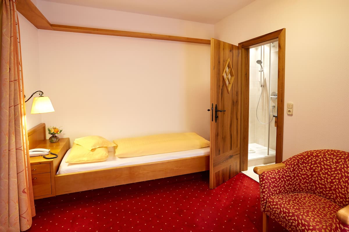 Une chambre d’hôtel avec un lit simple, une armoire en bois, un tapis rouge à pois, une chaise en bois et une salle de bain visible à travers une porte ouverte.