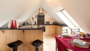 A kitchen with a central island, wooden cabinets, a microwave, and a toaster. There is a dining area with a table set for two, a red tablecloth, and a vase with a flower. - Pentre Riding Stable and Accommodation (Swansea)