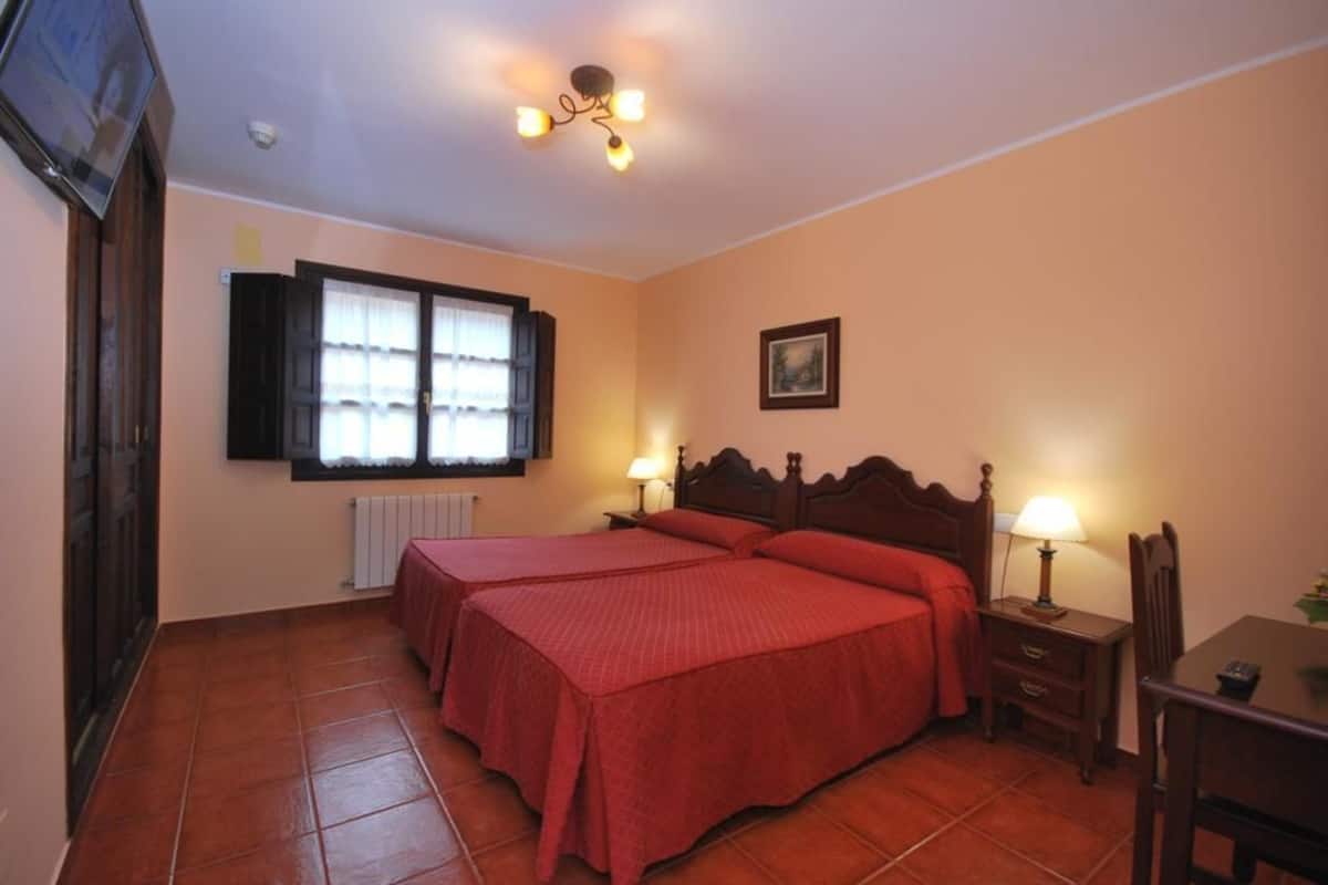 Un dormitorio con dos camas, una ventana con contraventanas, un radiador, cabecera de madera, lámparas de noche, una mesa y una silla.