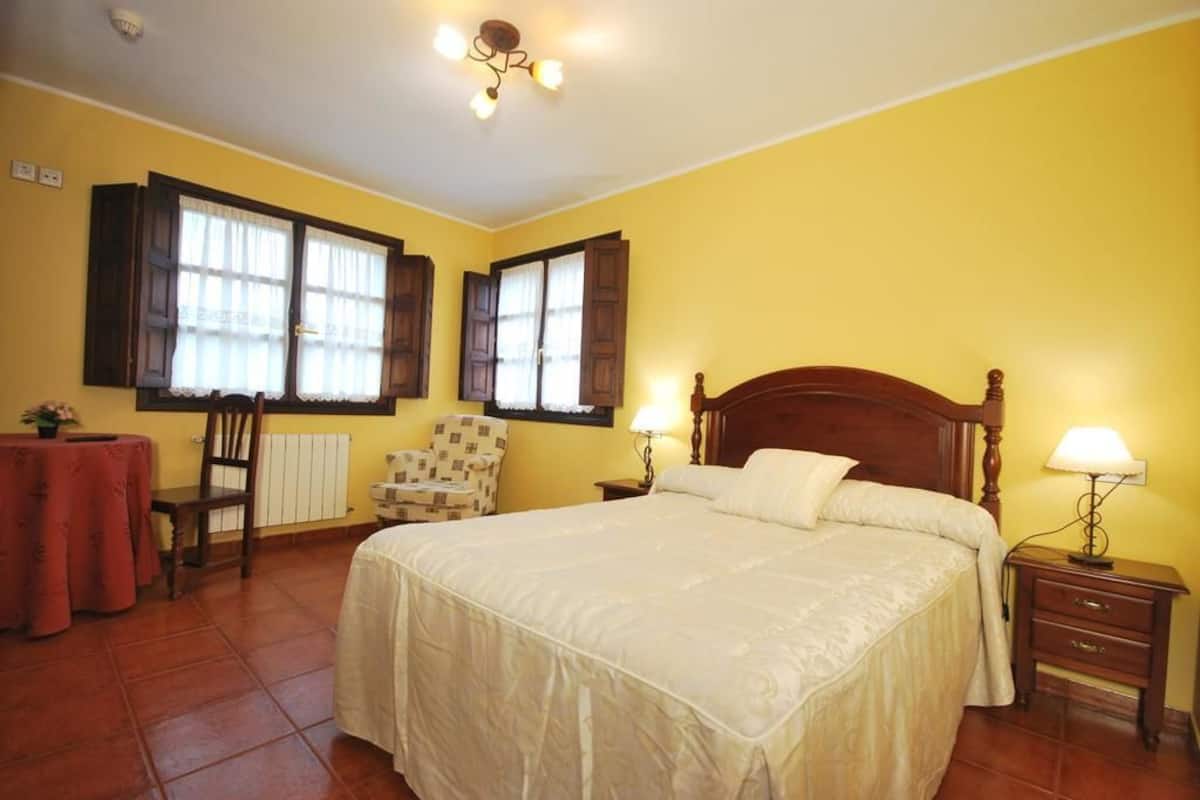 Un dormitorio con una cama grande, dos ventanas con cortinas, una silla, una mesita y una lámpara de noche.
