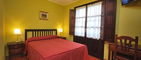 Una habitación con cama, dos mesitas de noche con lámparas, una silla de madera, televisor en la pared y una ventana con cortinas.