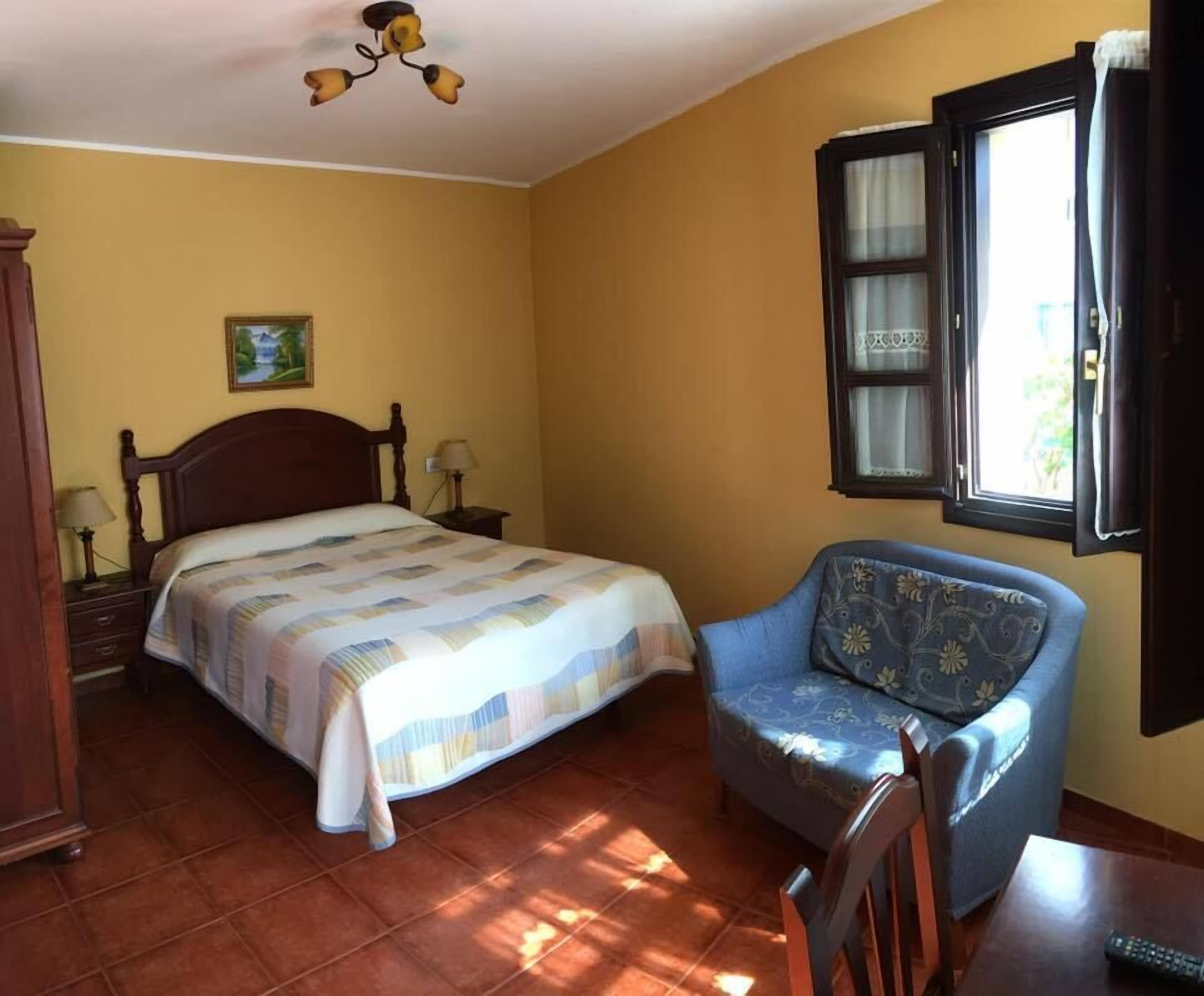 Un dormitorio con cama, una silla, una mesita de noche, lámpara, ventana y ventilador de techo.