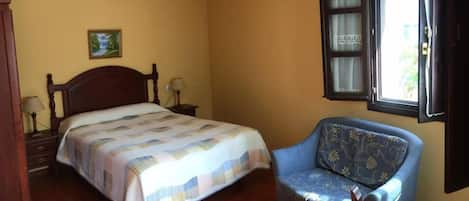 Un dormitorio con cama, una silla, una mesita de noche, lámpara, ventana y ventilador de techo.