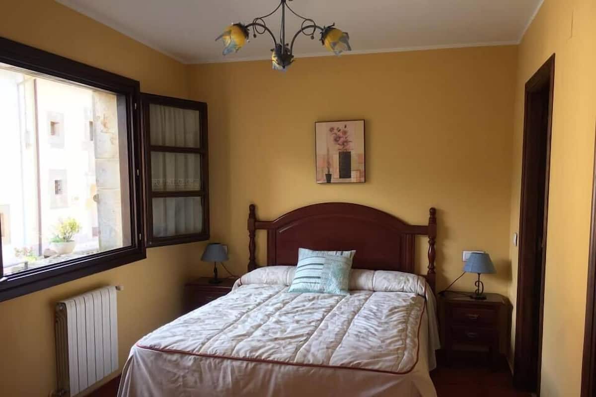 Un dormitorio con una cama, dos mesitas de noche con lámparas, una ventana con cortinas, un radiador y un cuadro colgado en la pared.