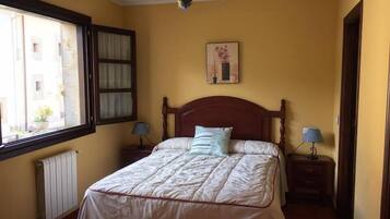 Quarto com cama, duas mesas de cabeceira com abajures, janela com cortinas, radiador e um quadro pendurado na parede.