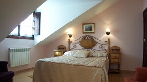 A bedroom with a sloped ceiling, a bed with a wooden headboard, two bedside tables with lamps, a radiator, and a window with curtains. - Hotel Los Ángeles (Cabrales)