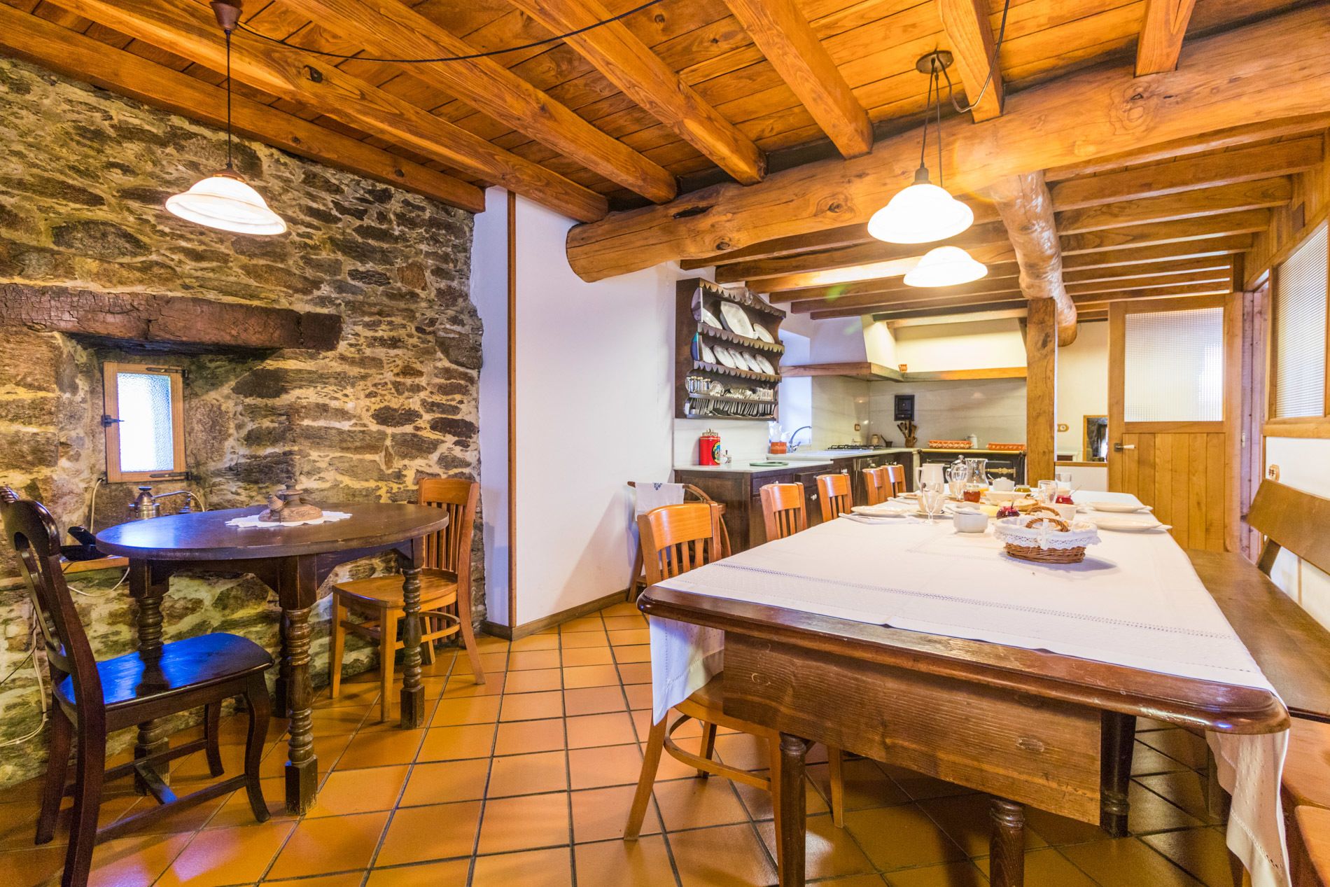 Un coin repas rustique avec une longue table en bois dressée pour un repas, entourée de chaises en bois. On peut apercevoir un mur en pierre et des poutres en bois au plafond, et un espace cuisine se trouve à l’arrière-plan.