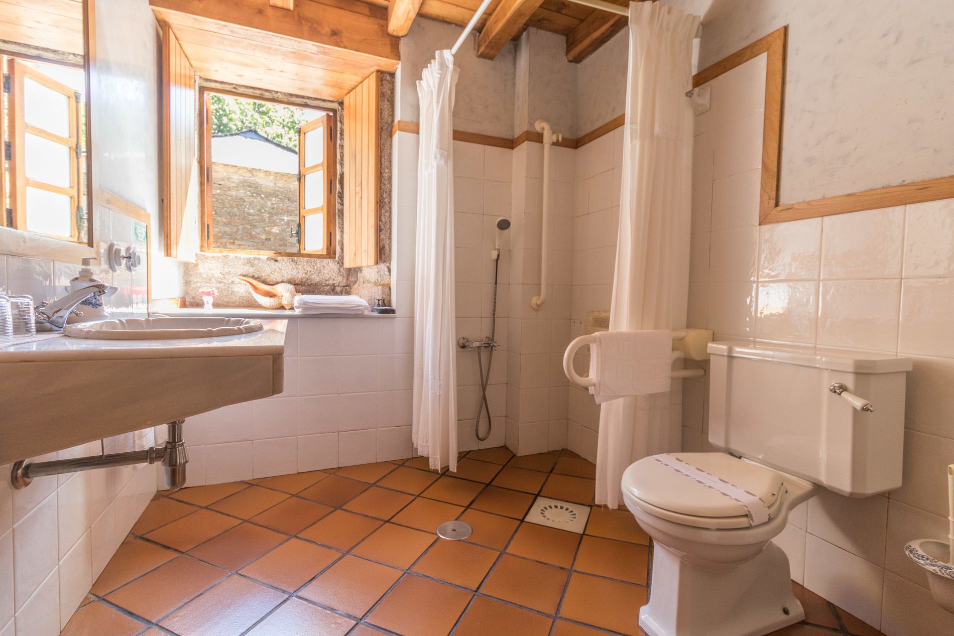 A bathroom with a sink, toilet, shower, and a window with wooden shutters.