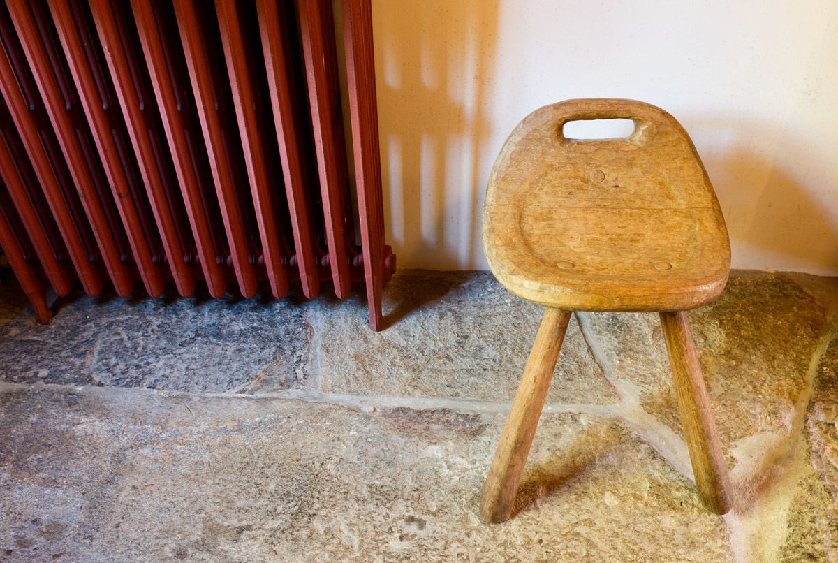 A wooden stool with a handle, placed on a stone floor next to a radiator.
