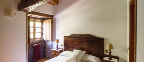 A bedroom with a wooden headboard, a bed with white linens, a nightstand with a lamp, and a window with a view of the outside.