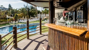 A poolside bar with a wooden counter, bar stools, and a view of the pool and surrounding greenery.