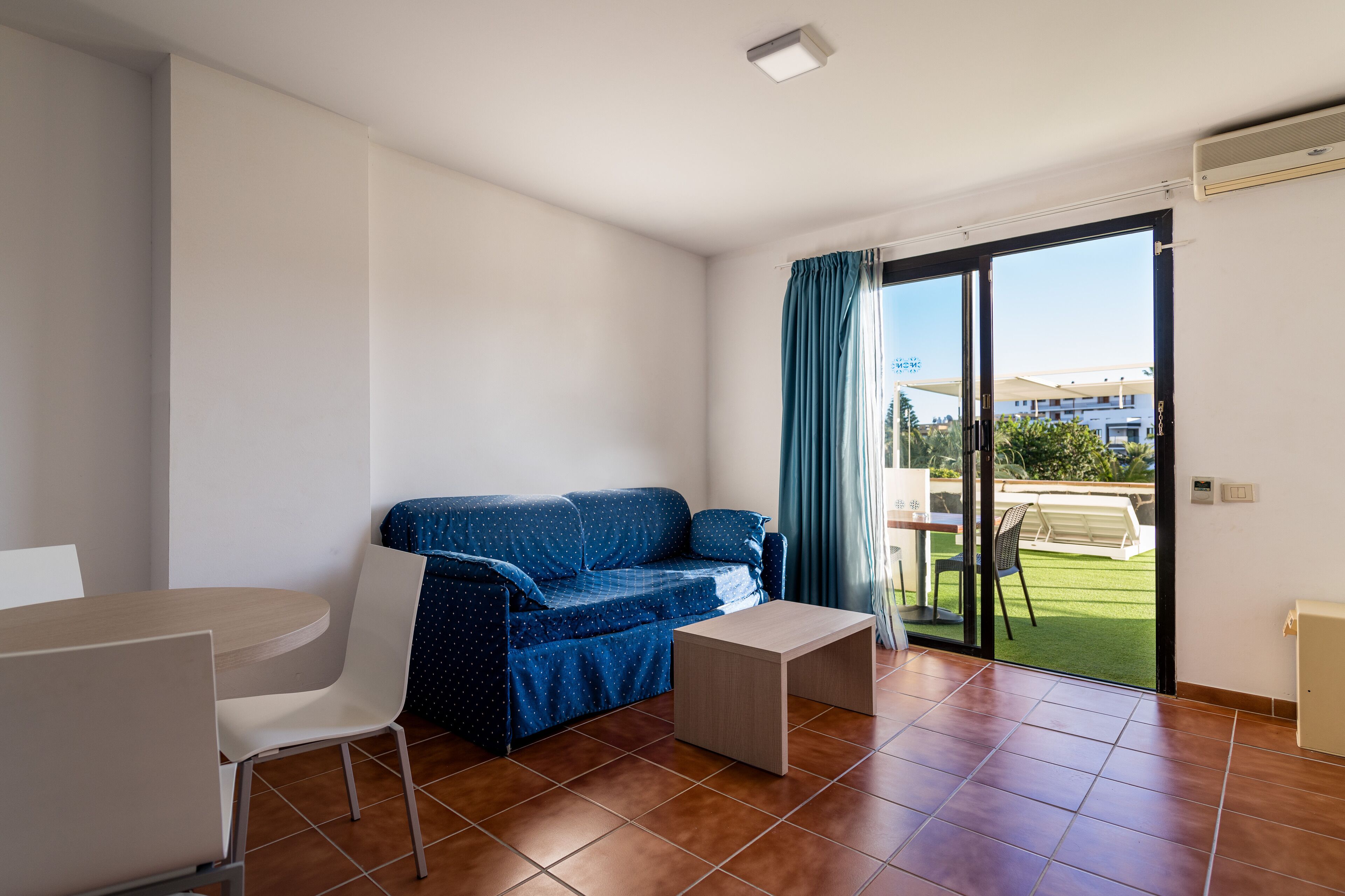 Un salón moderno con un sofá azul, una mesita y una silla. Hay un ventanal corredero que da a un balcón con vistas a edificios y zonas verdes.