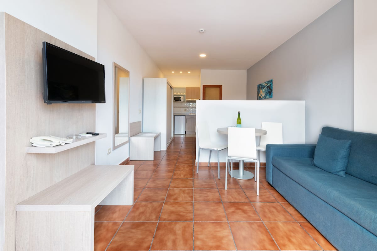 A modern hotel room with a flat-screen TV mounted on the wall, a sofa, a dining area with a table and chairs, and a kitchen in the background.