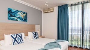 A hotel room with a large bed, two pillows, a painting on the wall, an air conditioner, and a view of the outdoors through curtains.