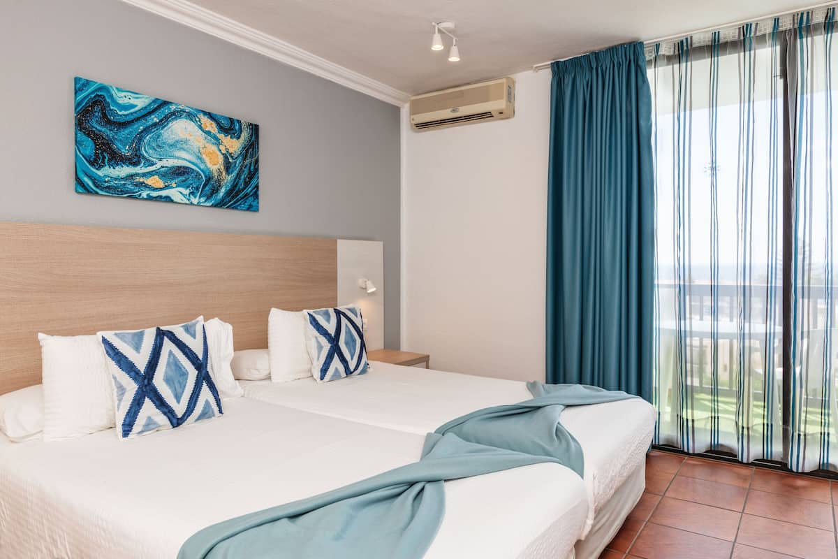 A hotel room with a large bed, two pillows, a painting on the wall, an air conditioner, and a view of the outdoors through curtains.