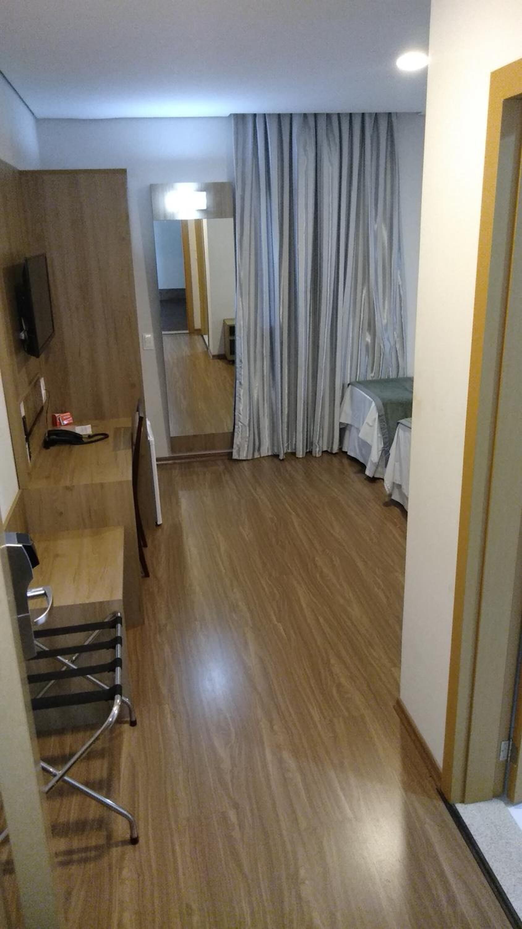 Une chambre d’hôtel avec un plancher en bois, un lit recouvert d’une courtepointe blanche, une petite table recouverte d’une nappe blanche, une chaise, un téléviseur fixé au mur et une grande fenêtre avec des rideaux.