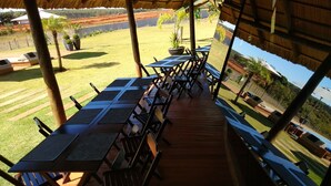 Un área de comedor al aire libre con varias mesas y sillas, techo de paja y vistas a un jardín ajardinado y una piscina.