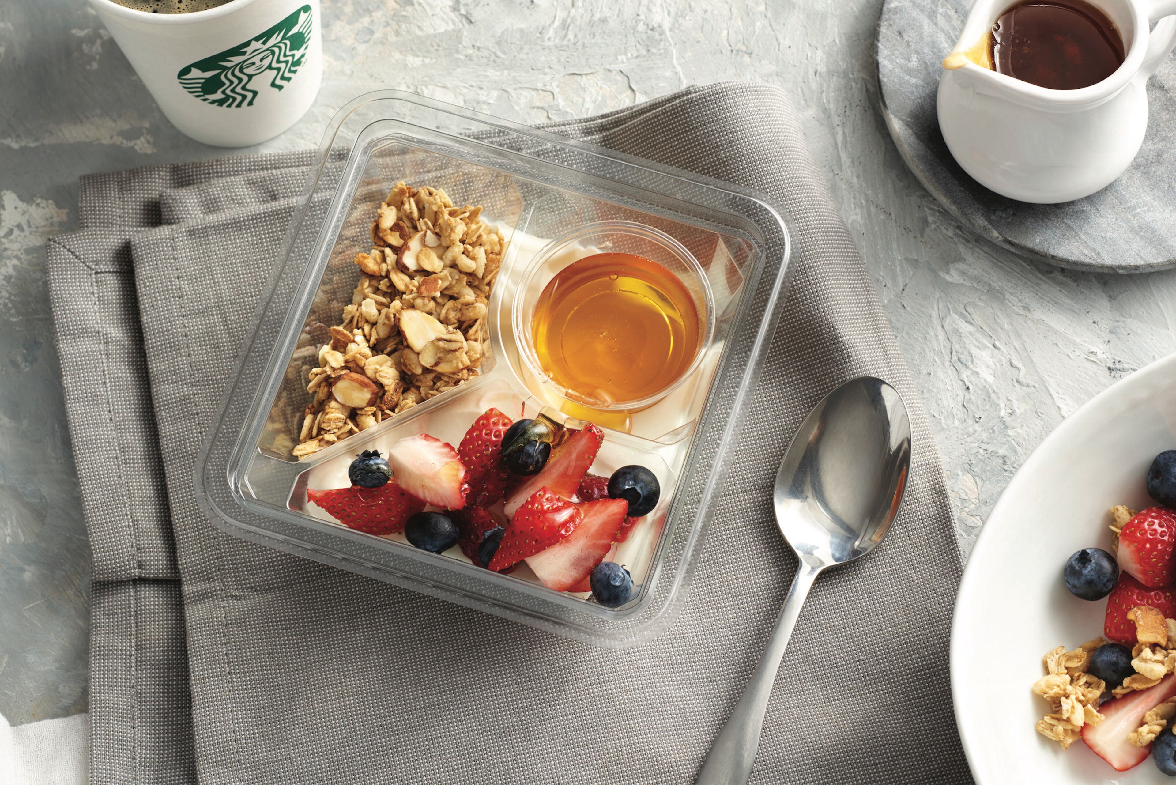 Un repas composé de granola, de yaourt et de baies, avec une cuillère et une tasse de café, le tout posé sur une table.
