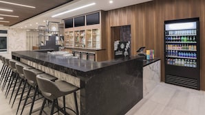 A modern bar area with a marble countertop, bar stools, a beverage vending machine, and a wine cooler. - Courtyard by Marriott Burlington (Burlington)