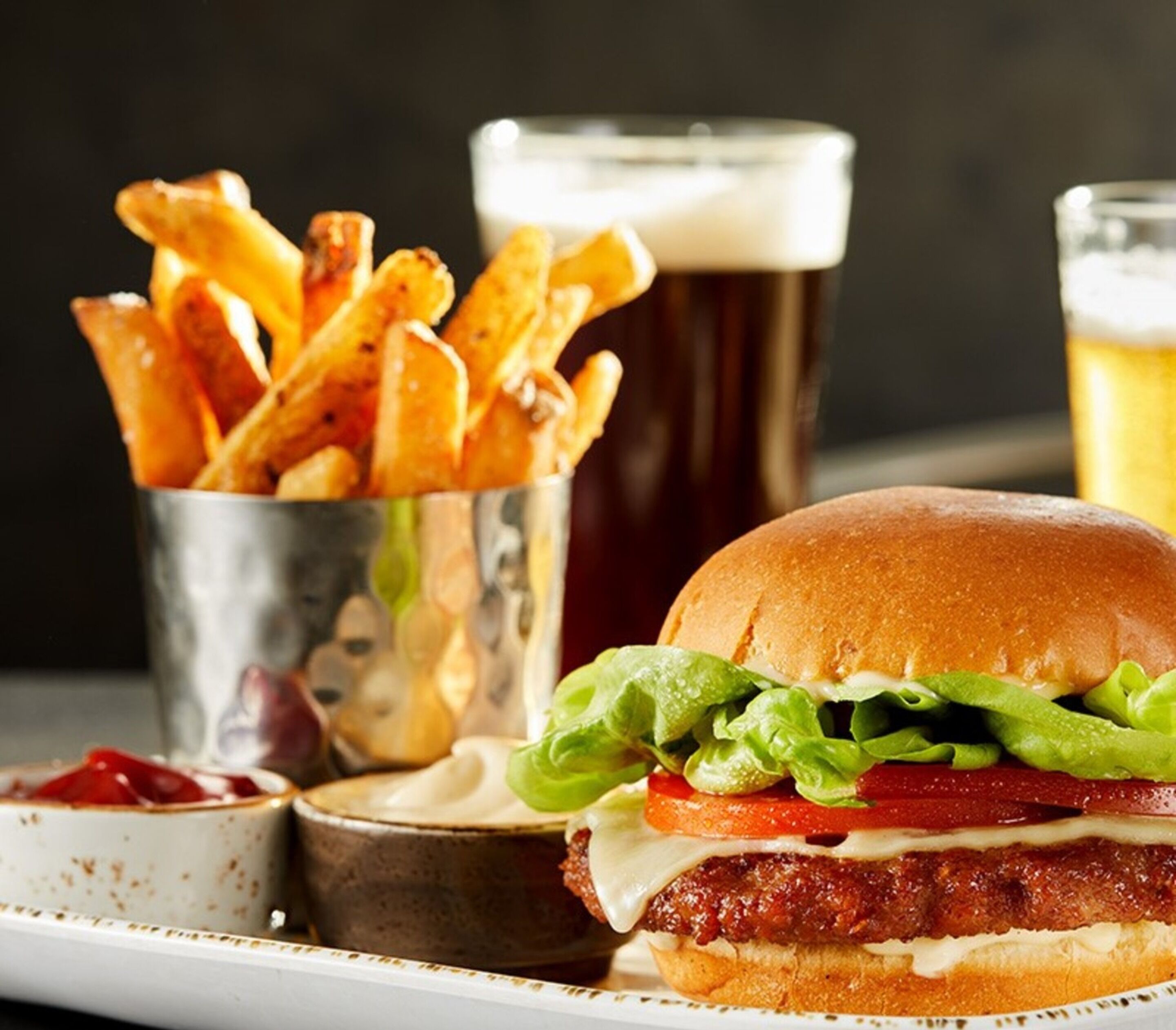 A burger with lettuce, tomato, and a bun, accompanied by a cup of fries and a glass of beer.