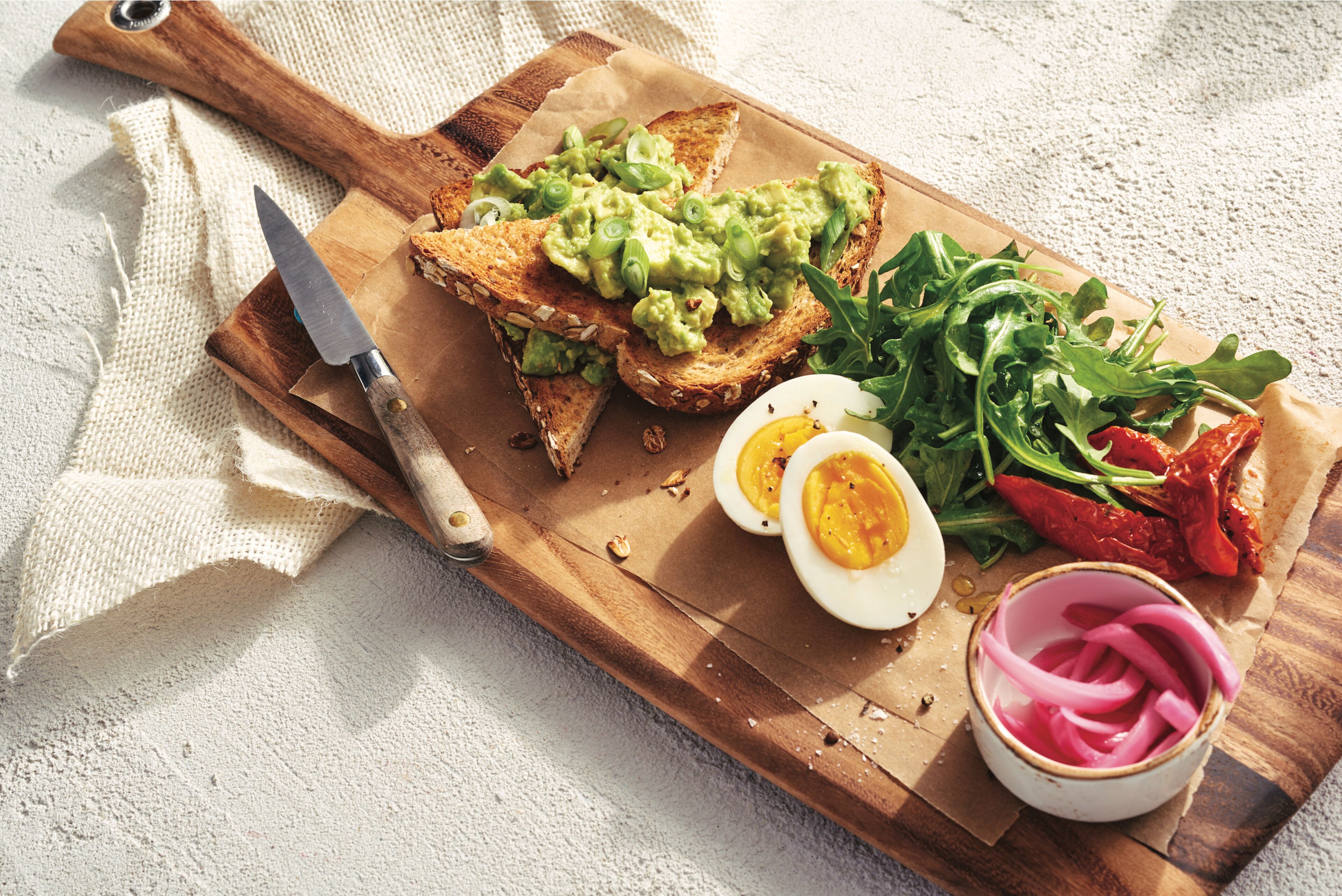 A wooden cutting board with toast topped with avocado, a halved boiled egg, arugula, and pickled red onions.