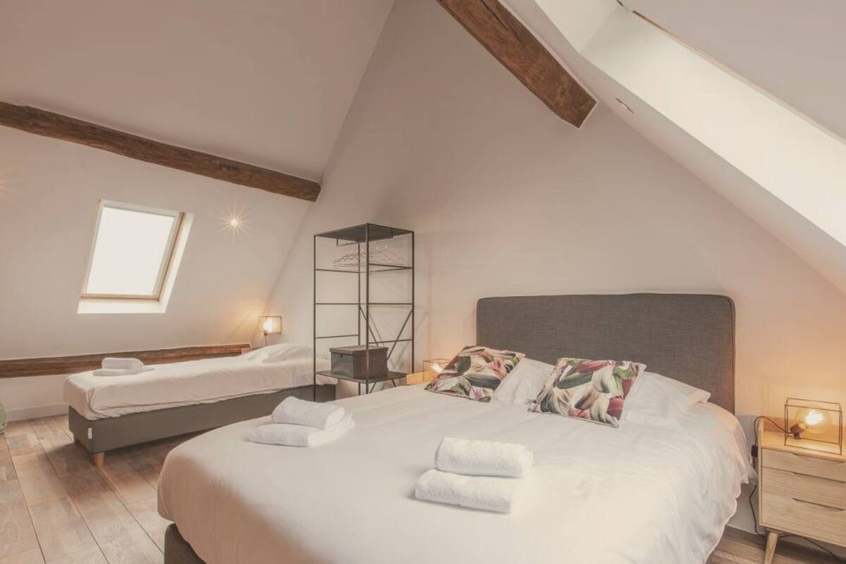 Une chambre avec un grand lit, un plus petit lit, une lucarne et une table de chevet avec une lampe.