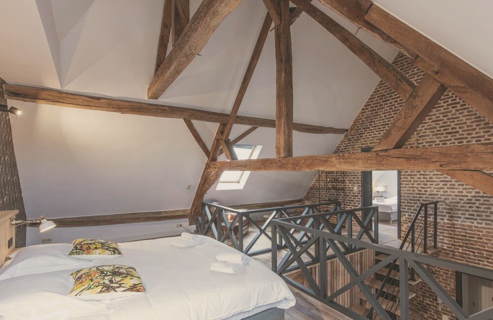 Un dormitorio con techo inclinado, vigas de madera a la vista, tragaluz y una cama con almohadas estampadas, además de vistas al baño.