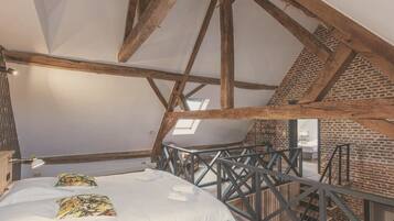 Un dormitorio con techo inclinado, vigas de madera a la vista, tragaluz y una cama con almohadas estampadas, además de vistas al baño.