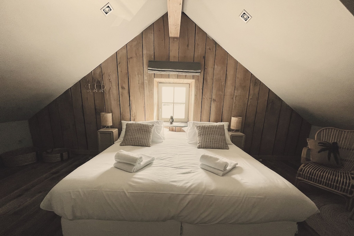 Une chambre avec un plafond en pente, des murs en bois, un grand lit avec du linge de lit blanc, deux tables de chevet avec des lampes et un fauteuil.