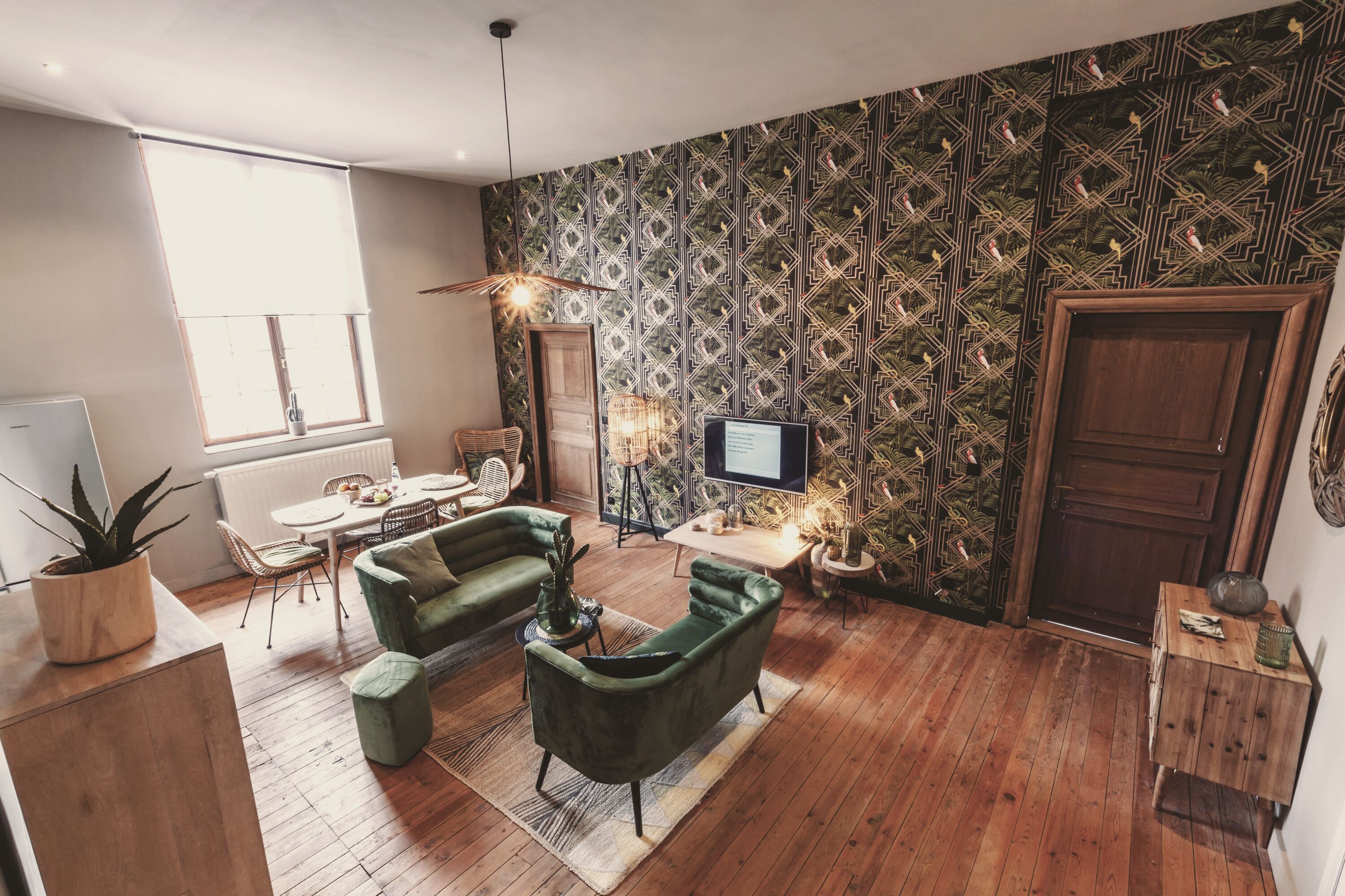 A modern living room with a green sofa, wooden flooring, a patterned wallpaper, a television, and a dining area with a table and chairs.