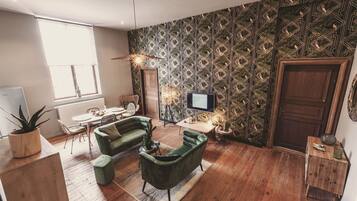 A modern living room with a green sofa, wooden flooring, a patterned wallpaper, a television, and a dining area with a table and chairs.