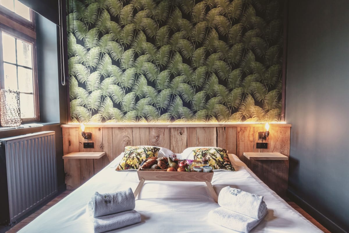Une chambre avec un lit, une tête de lit en bois, deux tables de chevet, un plateau avec des fruits et un ours en peluche, et une fenêtre donnant sur un beau paysage.