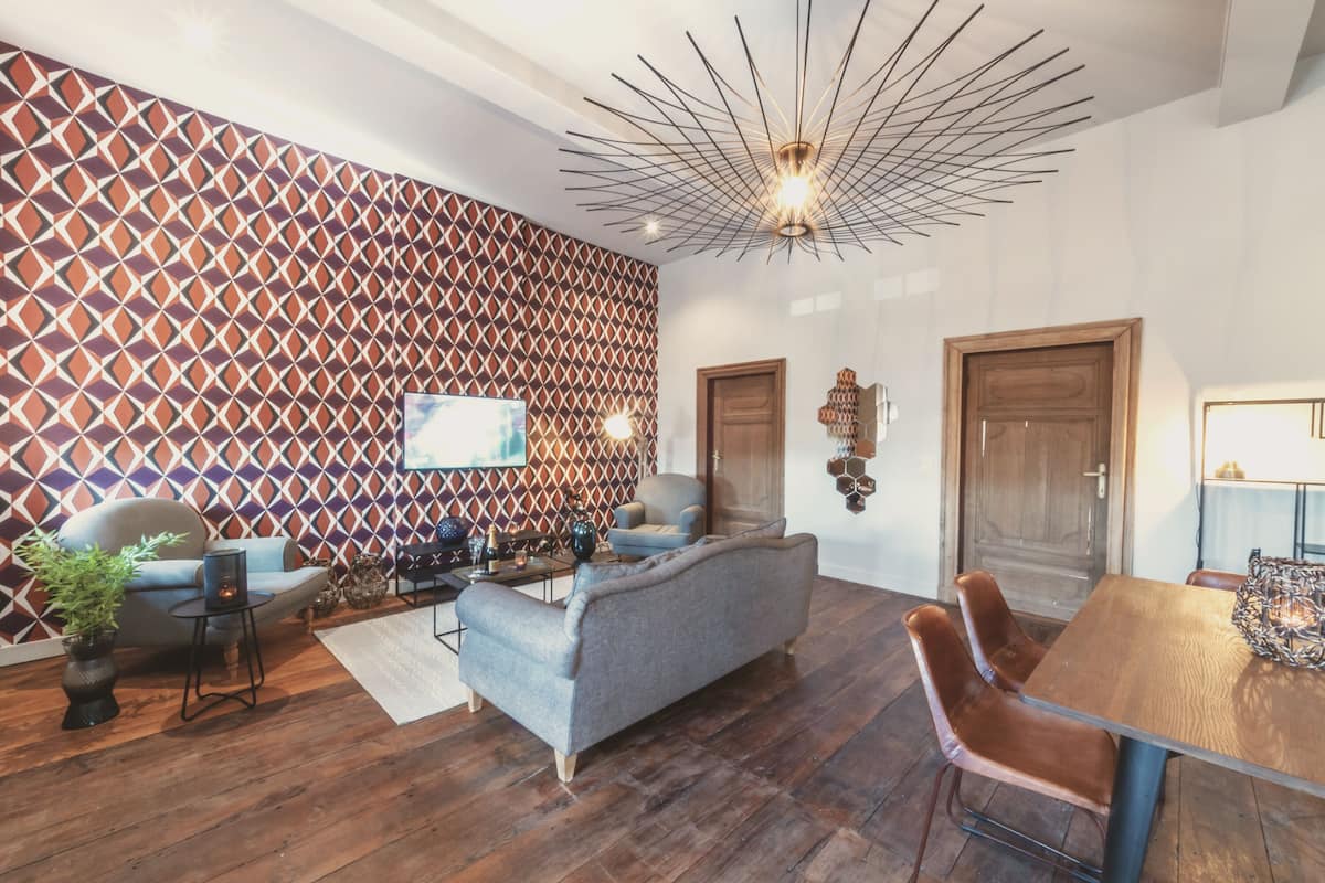 Un salon moderne avec un mur orné d’un motif géométrique, un canapé gris, une table à manger en bois avec des chaises, et un luminaire au plafond au design original.