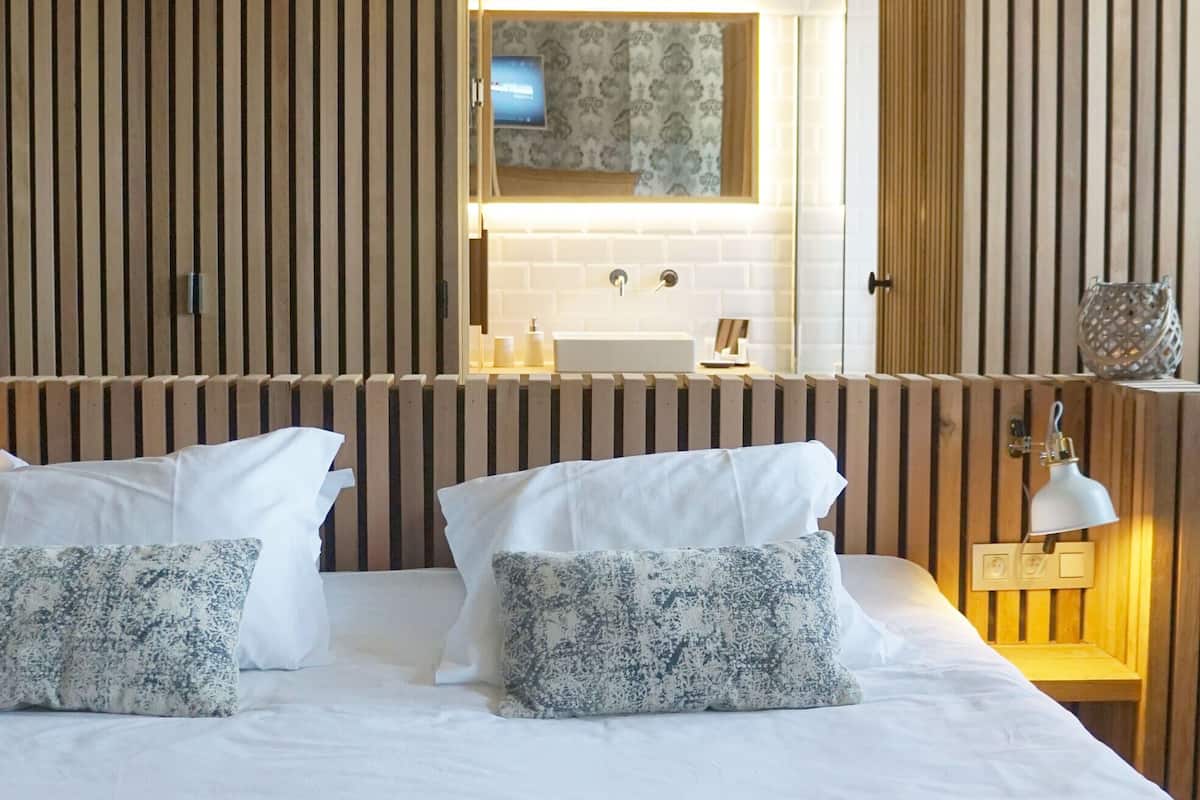 Une chambre d’hôtel avec un grand lit, deux oreillers, une table de chevet avec une lampe et un miroir au-dessus du lavabo.