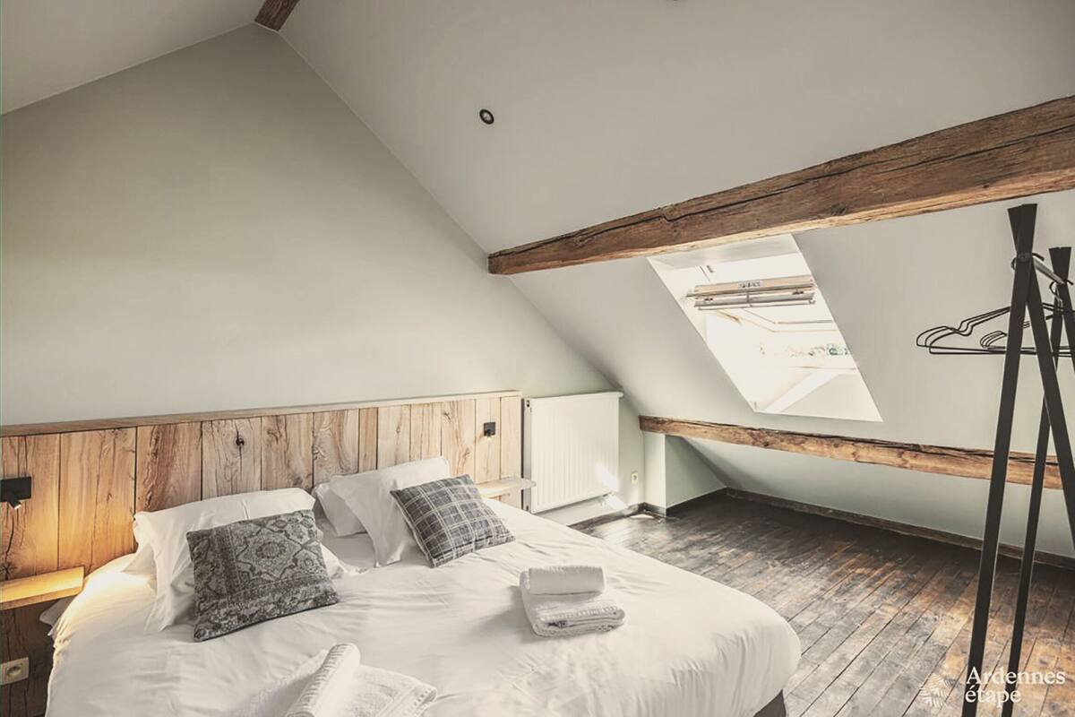 Une chambre avec un plafond en pente, des poutres en bois, un lit avec du linge de lit blanc, un puits de lumière et un radiateur.
