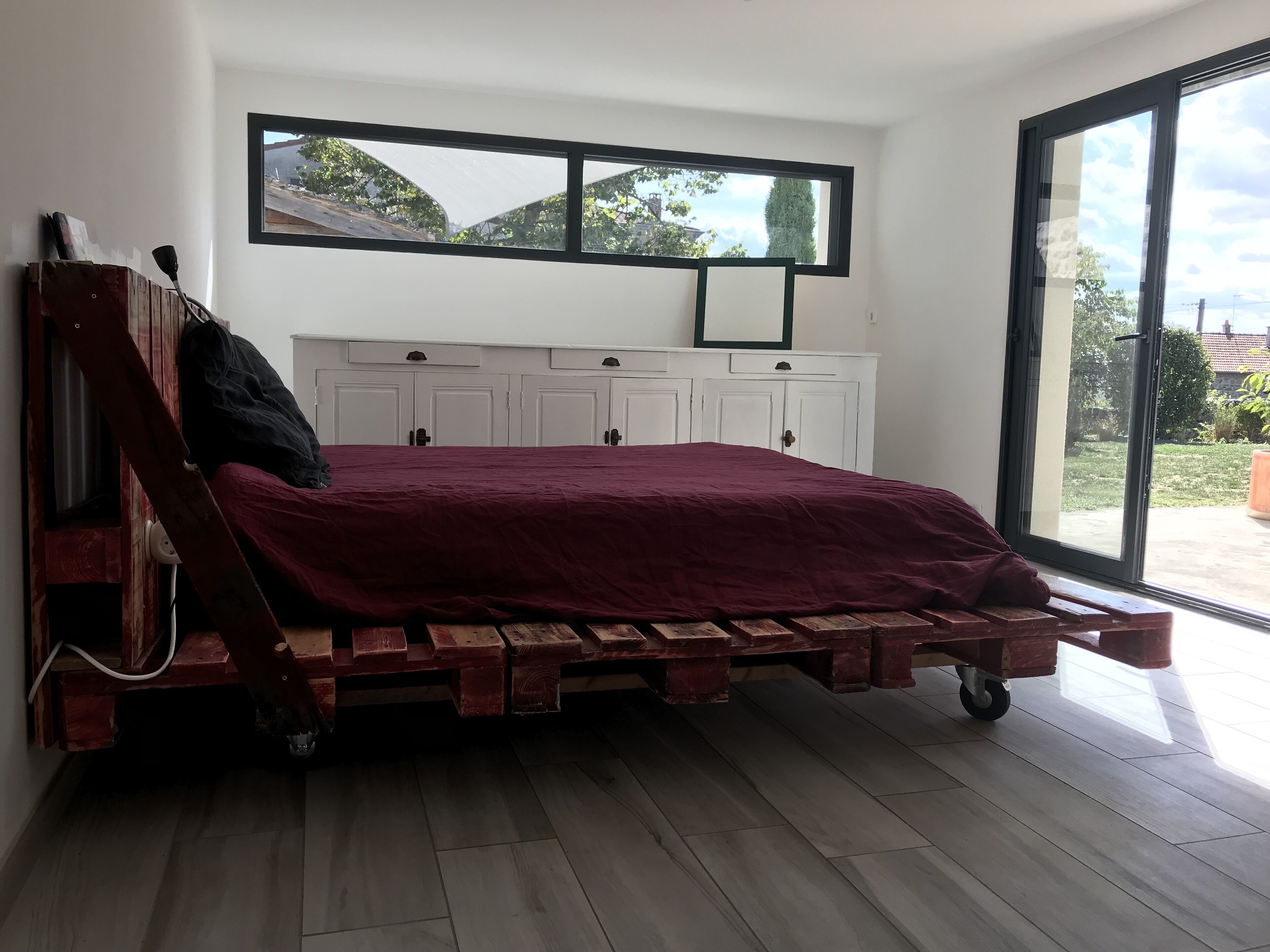 Een slaapkamer met een houten bedframe, een rode spreidek en een groot raam met uitzicht op de omgeving.