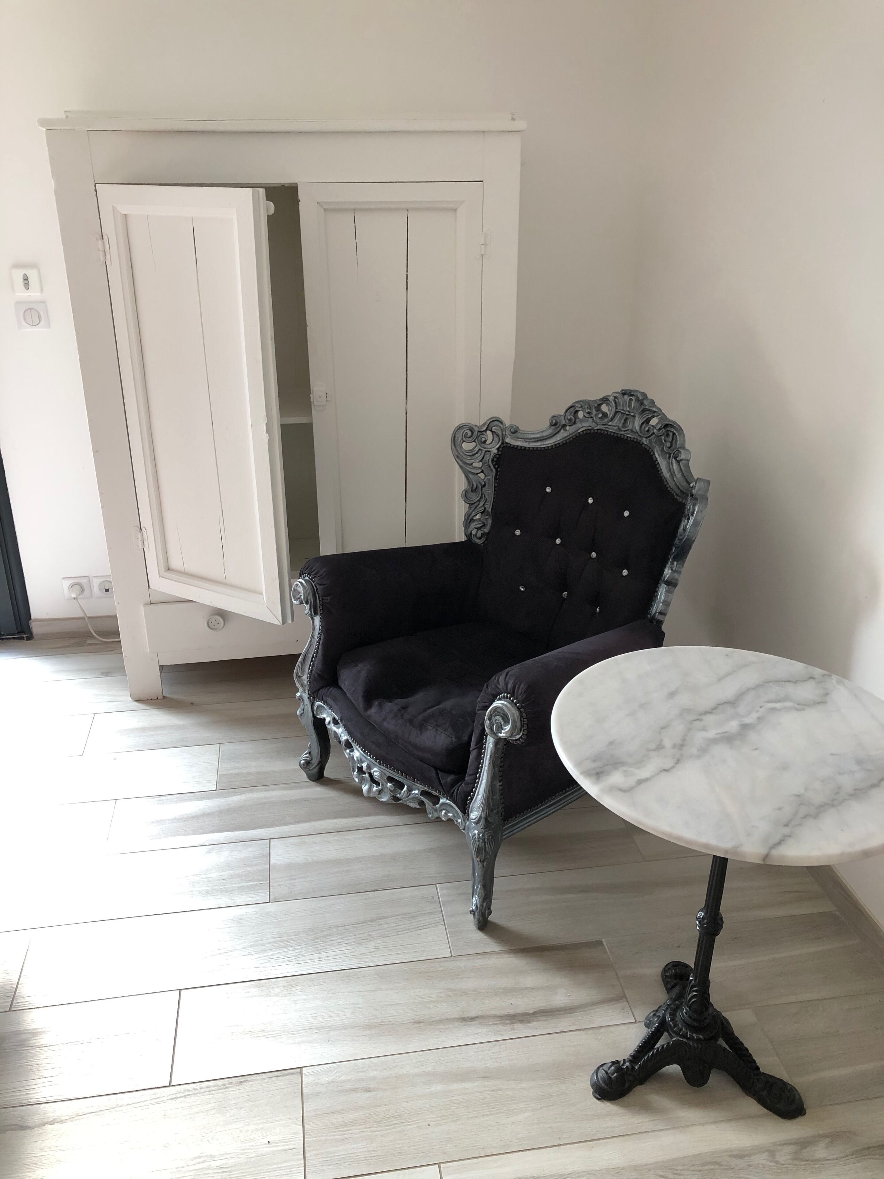 A room with a black upholstered chair with ornate detailing, a marble table, and a white wardrobe.