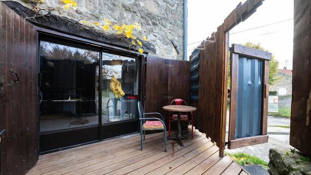 Eine Holzterrasse mit Glastür zu einem Raum, ein kleiner Tisch mit zwei Stühlen und eine Steinwand mit gelben Blumen.