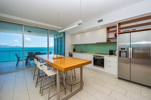 Eine moderne Küche mit einem großen, edlen Kühlschrank aus Edelstahl, einem Essbereich mit Holztisch und weißen Stühlen sowie Blick auf das Meer durch große Glasüren.