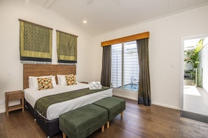 A bedroom with a large bed, a bench, a small table, and a window with curtains. - Reflections Amber Villa 2 (Picnic Bay)