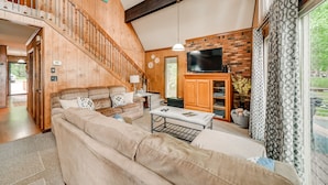 A spacious living room with a high ceiling, wooden staircase, a flat-screen TV, and a large window with patterned curtains.