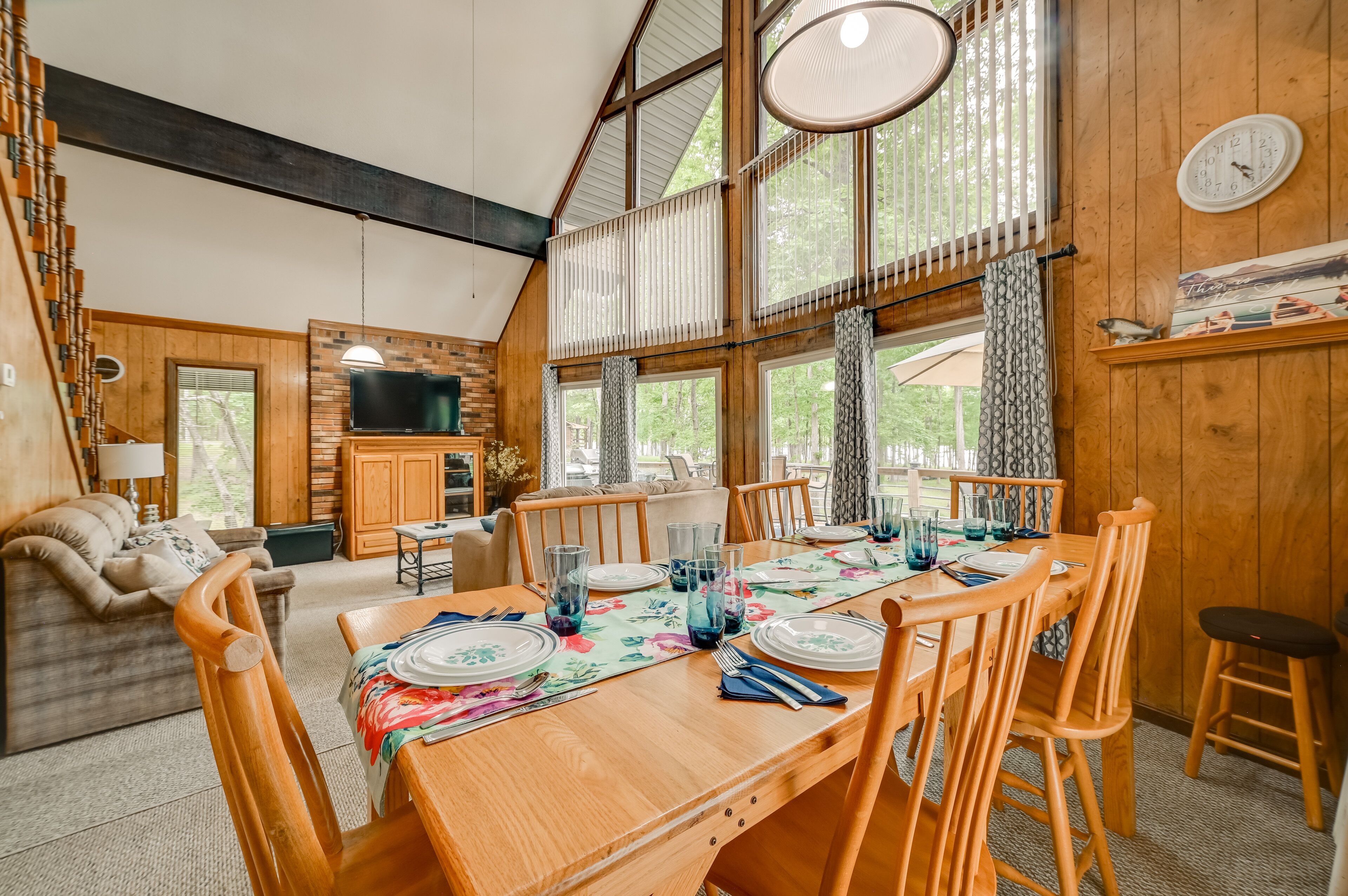 L’intérieur d’un chalet accueillant, avec un coin repas, des murs en bois, de grandes fenêtres, un téléviseur et une horloge au mur.