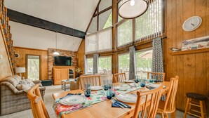 A cozy cabin interior with a dining area, wooden walls, large windows, a television, and a clock on the wall.