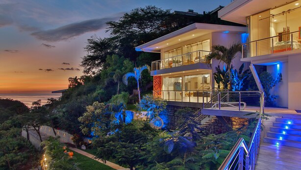 A modern multi-story house with a balcony, surrounded by lush greenery and a view of the ocean at dusk. - The Malinche Palace (Sardinal)